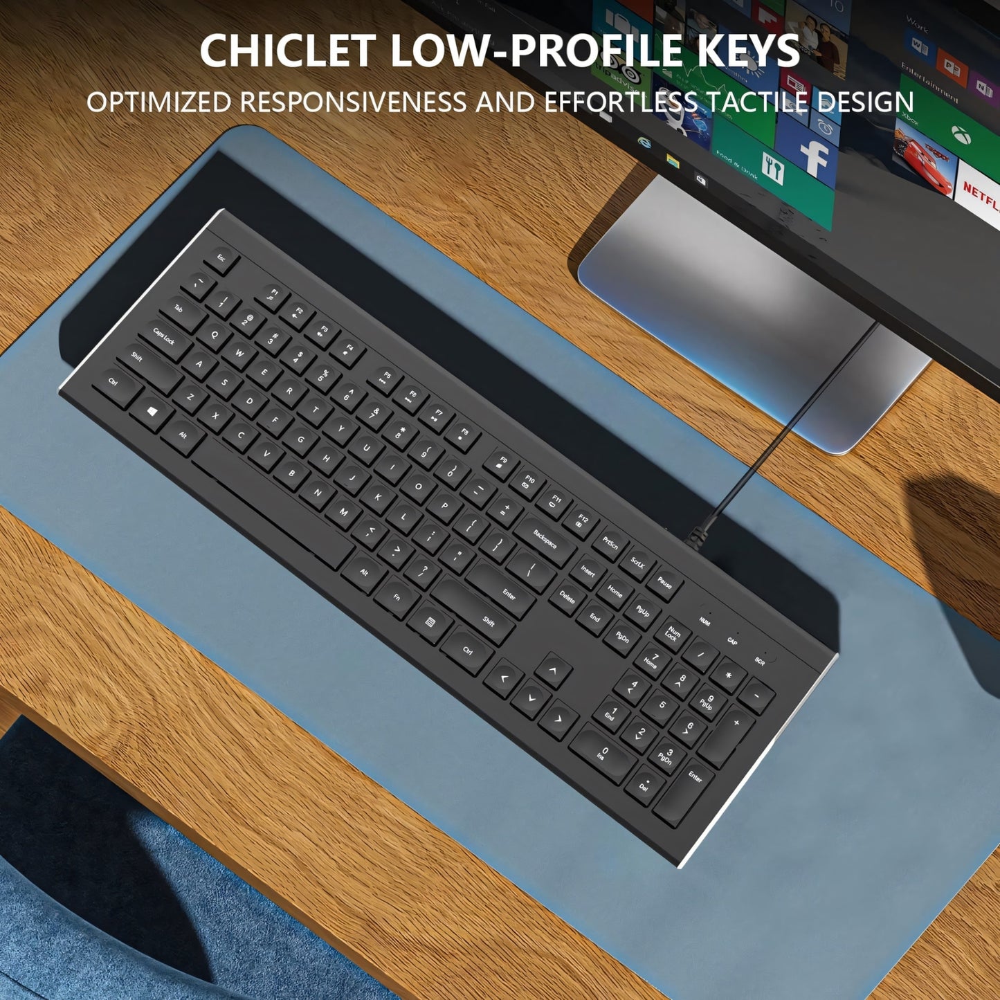 A modern and sleek workspace featuring an Clevisco ergonomic keyboard with chiclet low-profile keys, optimized for responsiveness and effortless tactile design. The keyboard is placed on a stylish blue desk mat. In the background, a monitor displaying various app icons is visible on the polished wooden desk. A person’s knee is partially visible at the bottom of the image, indicating that they are sitting at the desk